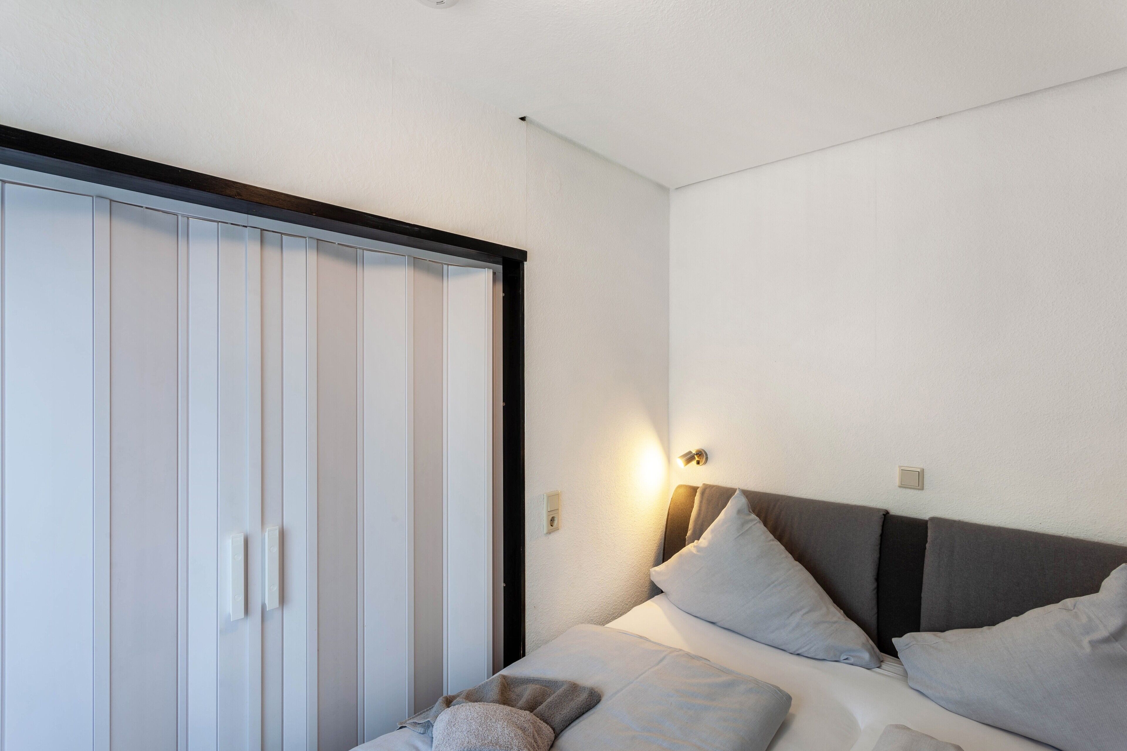 Un dormitorio con cama, almohadas, mesita de noche, lámpara de pared y armario.