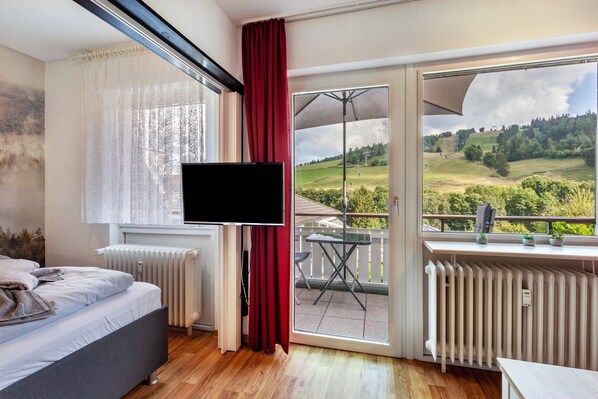 A bedroom with a bed, a TV, a radiator, and a balcony with a table and chairs.