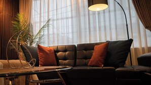 Un salon moderne avec un canapé d’angle, une lampe sur pied et une grande fenêtre ornée de rideaux légers.