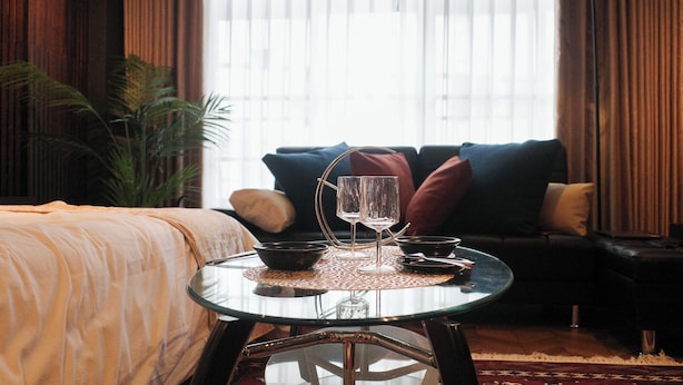 Une chambre d’hôtel avec une table en verre sur laquelle sont disposés un bol, deux verres à vin et une bougie. On y trouve également un canapé avec des coussins et une grande fenêtre avec des rideaux.