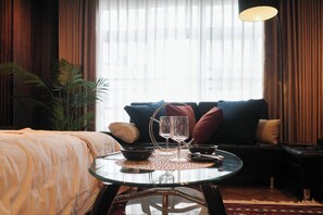 Une chambre d’hôtel avec une table en verre sur laquelle sont disposés un bol, deux verres à vin et une bougie. On y trouve également un canapé avec des coussins et une grande fenêtre avec des rideaux.