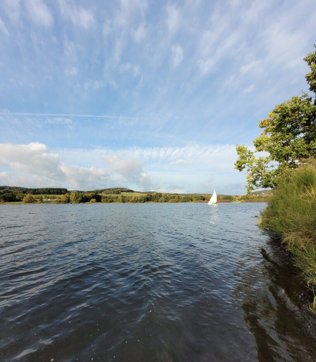 Ein ruhiger See mit einem Segelboot, umgeben von viel Grün und Hügeln.