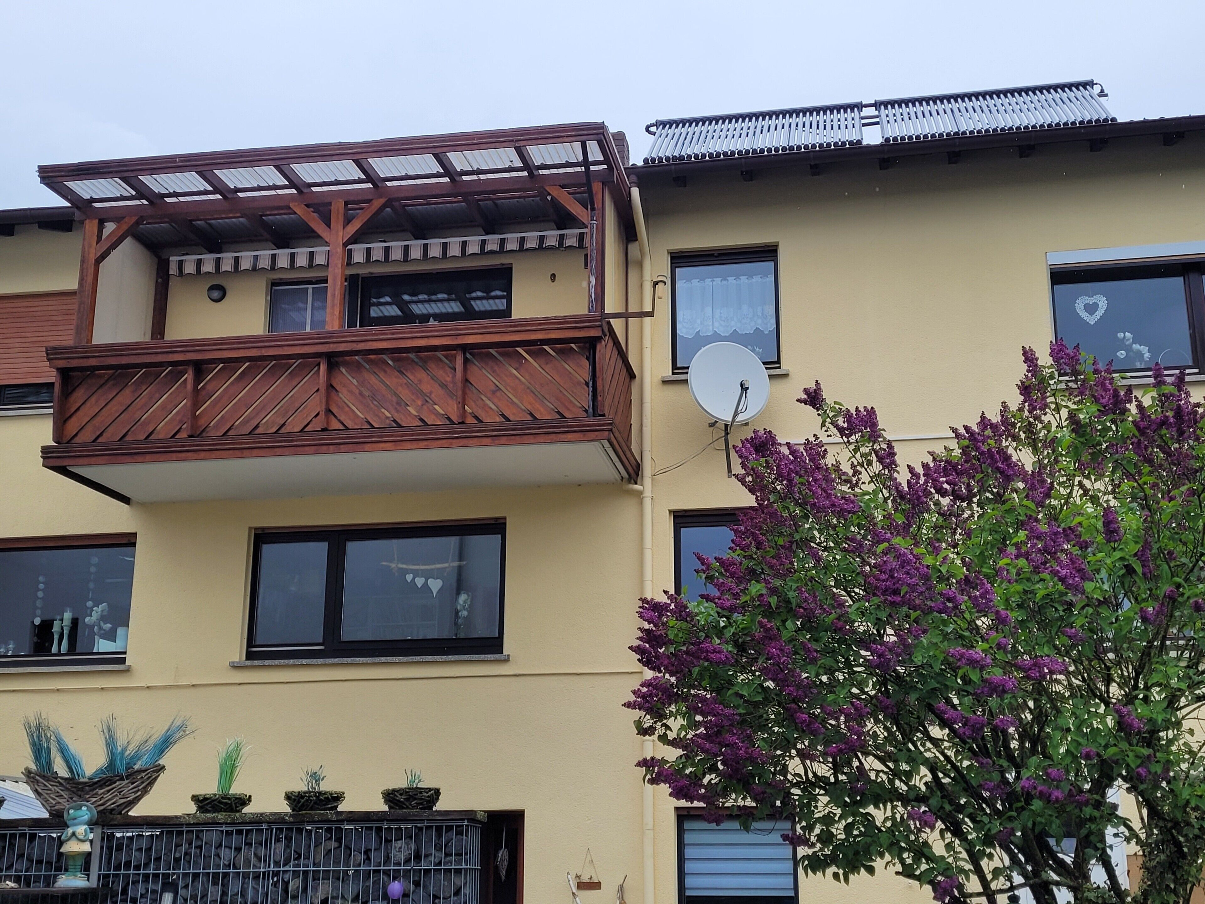 Ein zweistöckiges Gebäude mit Balkon, Satellitenschüssel und einem Baum mit violetten Blüten.