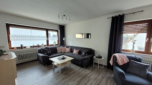 A living room with a sectional sofa, a coffee table, a TV mounted on the wall, and a window with blinds. - Holiday Apartment "Am Strohberg" with Balcony and Wi-Fi (Gladenbach/ Weidenhausen)