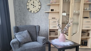 A room with a grey armchair, a black table with a pink vase, a wall clock, and a wooden cabinet with glassware. - Holiday Apartment "Am Strohberg" with Balcony and Wi-Fi (Gladenbach/ Weidenhausen)