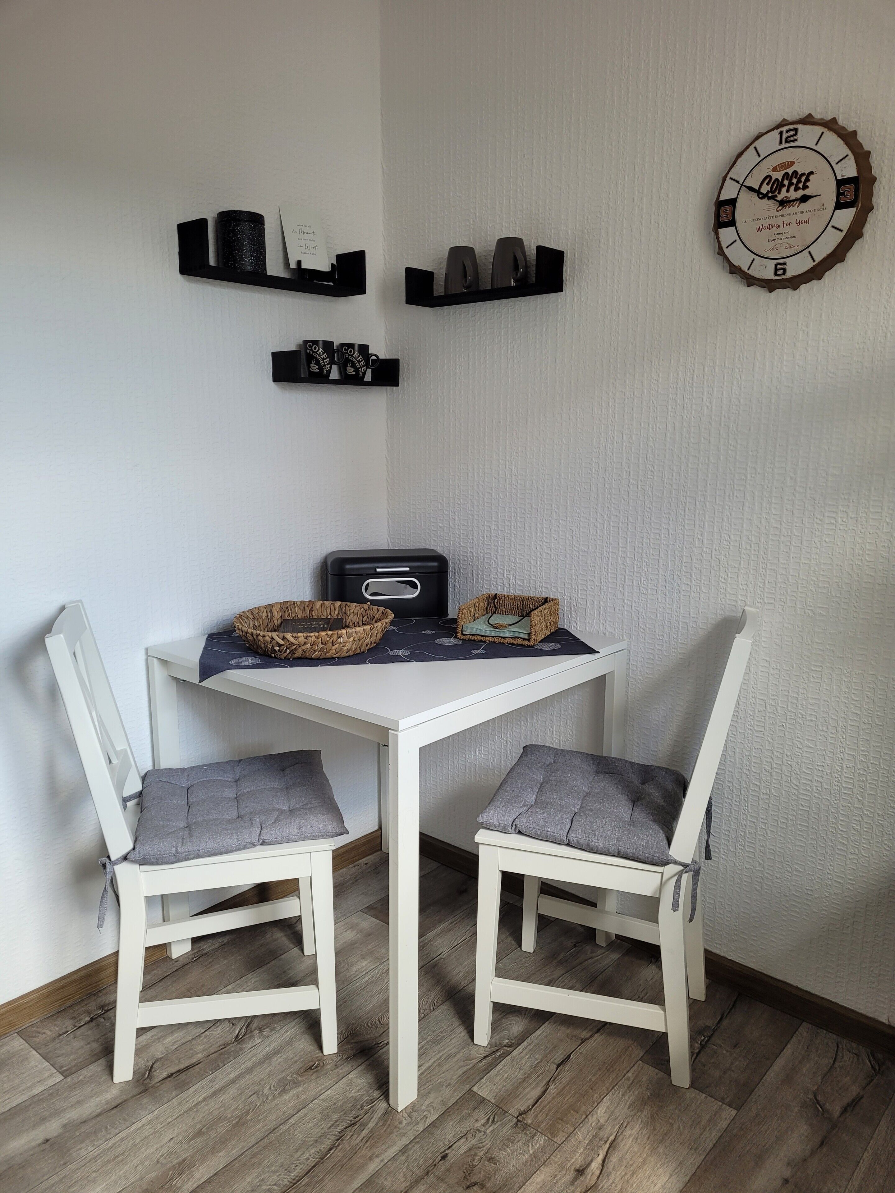 A small dining area with a white table and two chairs, a coffee machine, and shelves with coffee mugs.