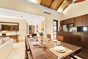 Une salle à manger moderne avec une table en bois dressée pour un repas, un coin cuisine avec des meubles de couleur bois foncé et un ventilateur de plafond.