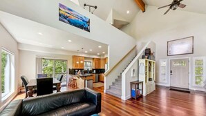 A spacious living area with a black leather sofa, a blue ottoman, wooden flooring, a kitchen in the background, and a staircase leading to an upper level. - Forest Mtn Lodge: Hot Tub, Game Room, Near Trail (South Lake Tahoe)