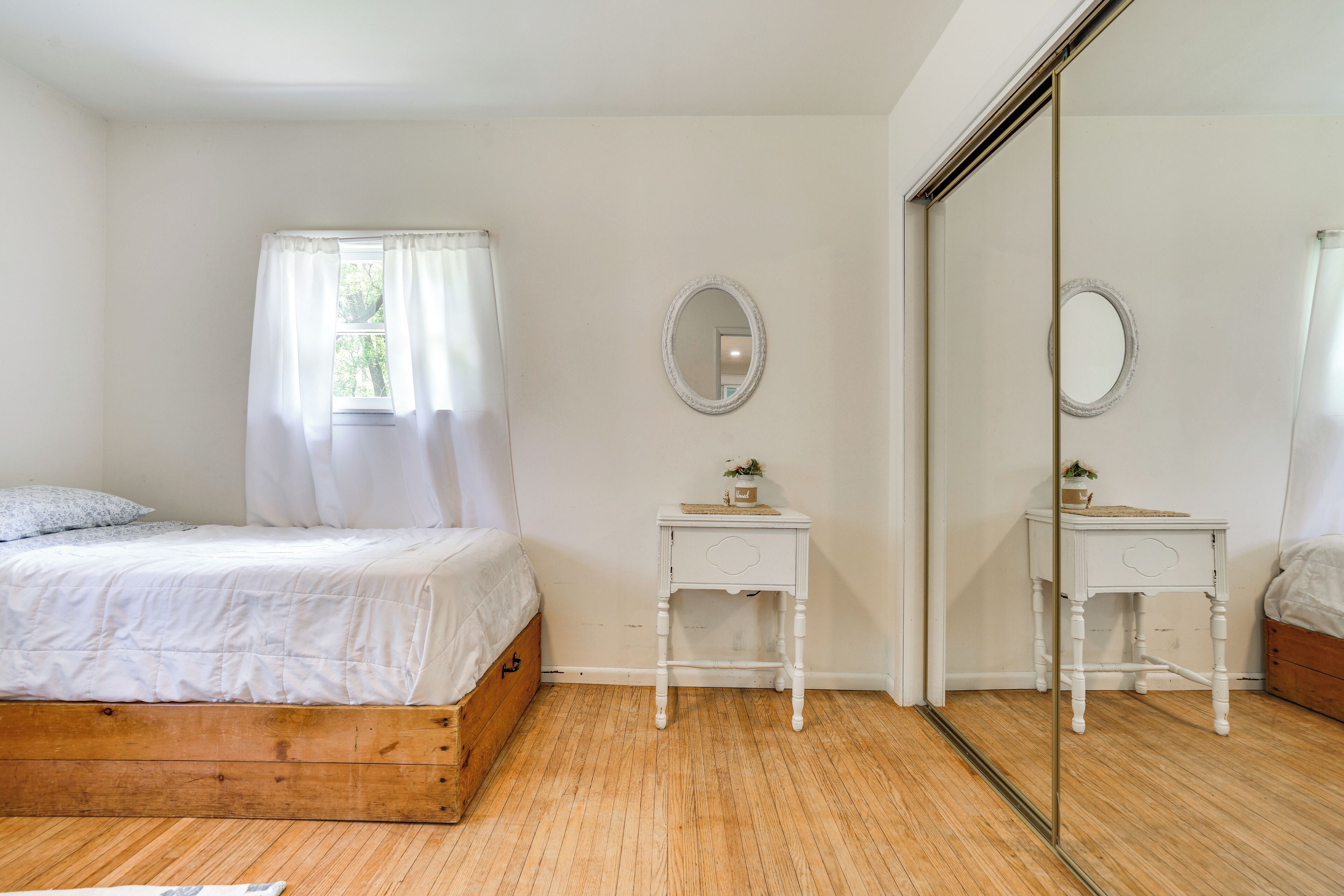 Quarto com cama de madeira, cortinas brancas, mesa de cabeceira com espelho e guarda-roupa.