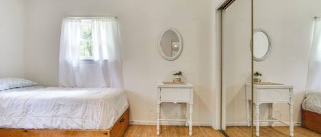 Quarto com cama de madeira, cortinas brancas, mesa de cabeceira com espelho e guarda-roupa.