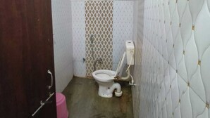 A small, compact bathroom with a toilet, a wooden door, and a pink bucket. - Hotel Basant Palace (Gopadbanas)