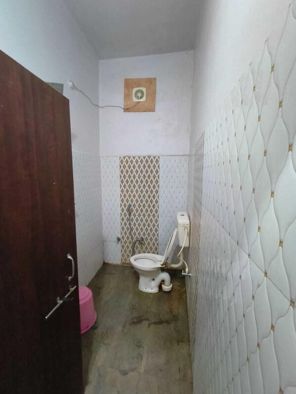 A small, compact bathroom with a toilet, a wooden door, and a pink bucket. - Hotel Basant Palace (Gopadbanas)
