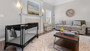 Ein Wohnzimmer mit einem schwarzen Reisebett für Babys, einem grauen Sofa, einem Holztisch und einem gemusterten Teppich.