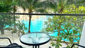 En balkon med et glasbord og to stole, der ud til et swimmingpool omgivet af tropiske planter.