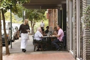Eine Restaurantterrasse mit Außenbestuhlung, ein Kellner kommt an einen Tisch, und eine Ziegelhausfassade.