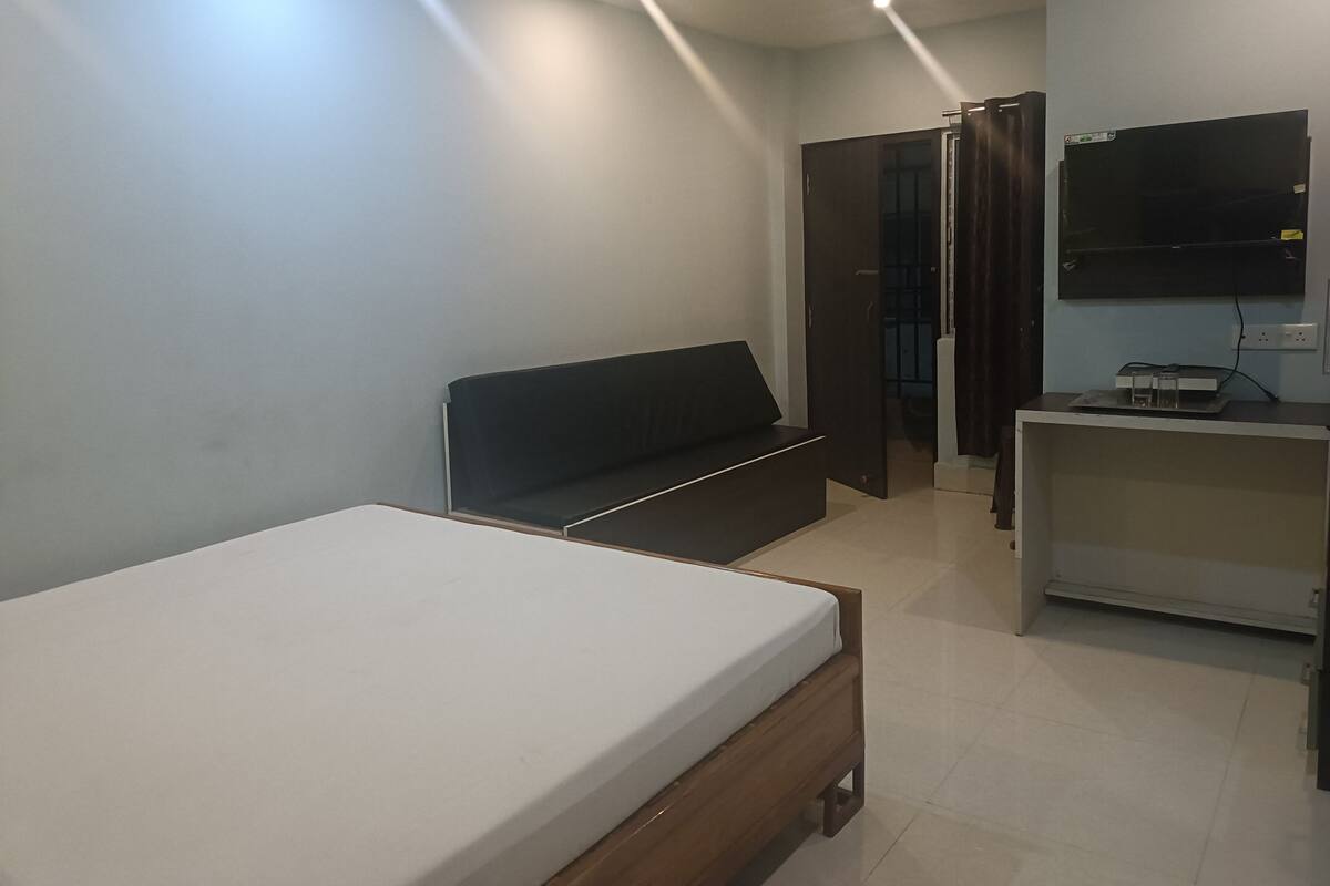 Un dormitorio moderno con cama, un sofá oscuro, un televisor montado en la pared y un escritorio pequeño con ordenador.