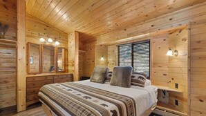 Een slaapkamer in een houten blokhut, met een bed, een dressoir, een spiegel en een raam met jaloezieën.