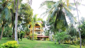 A two-story building with a red roof, surrounded by lush greenery and palm trees. - Peacock Point - Whole Villa 2 Bedroom (Sleeps 4) (Ahangama)