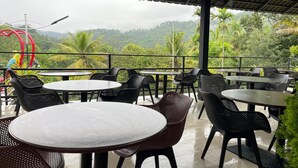 Un área de estar al aire libre con mesas y sillas redondas, con vistas a un paisaje exuberante con colinas y palmeras.