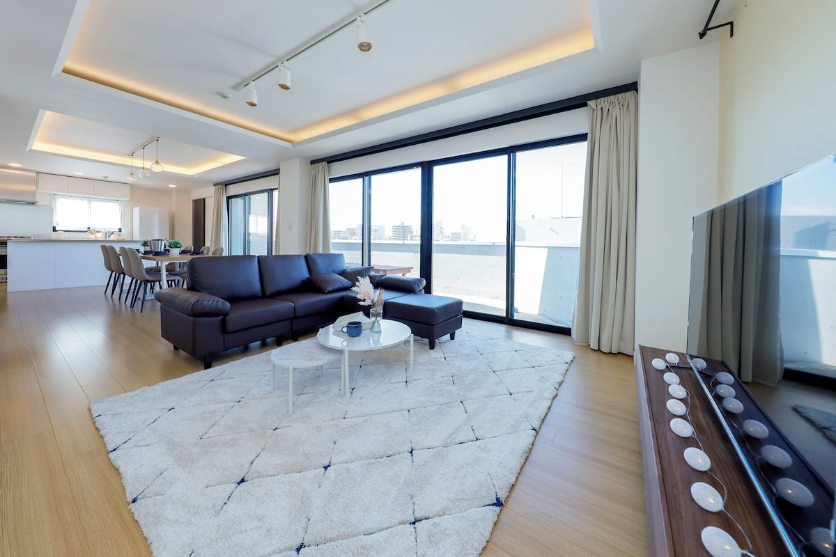 A modern living room with a sofa, coffee table, and a large rug. There is a dining area in the background with a table and chairs.