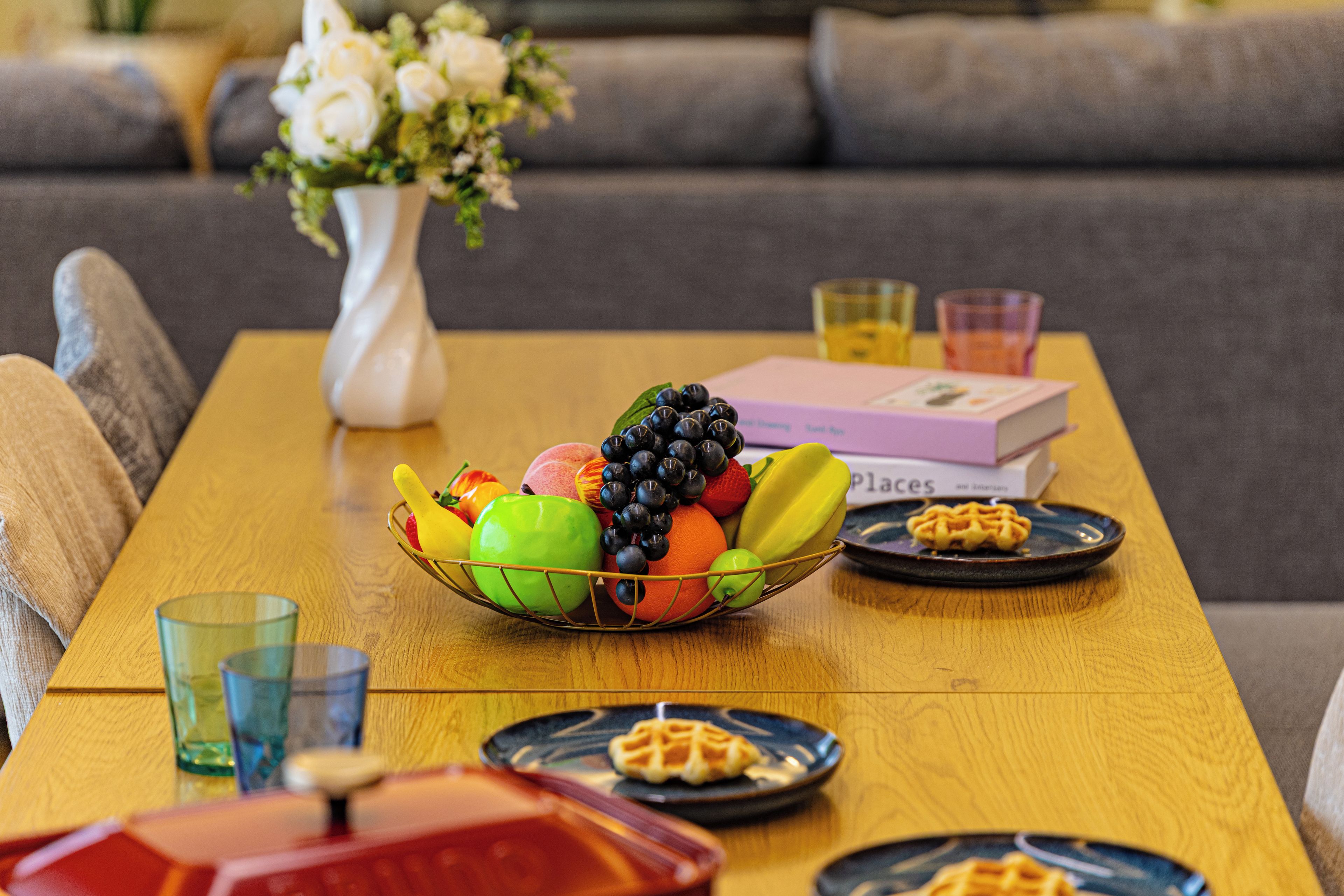 A dining table with a fruit bowl, two plates with waffles, a vase with flowers, and a book.