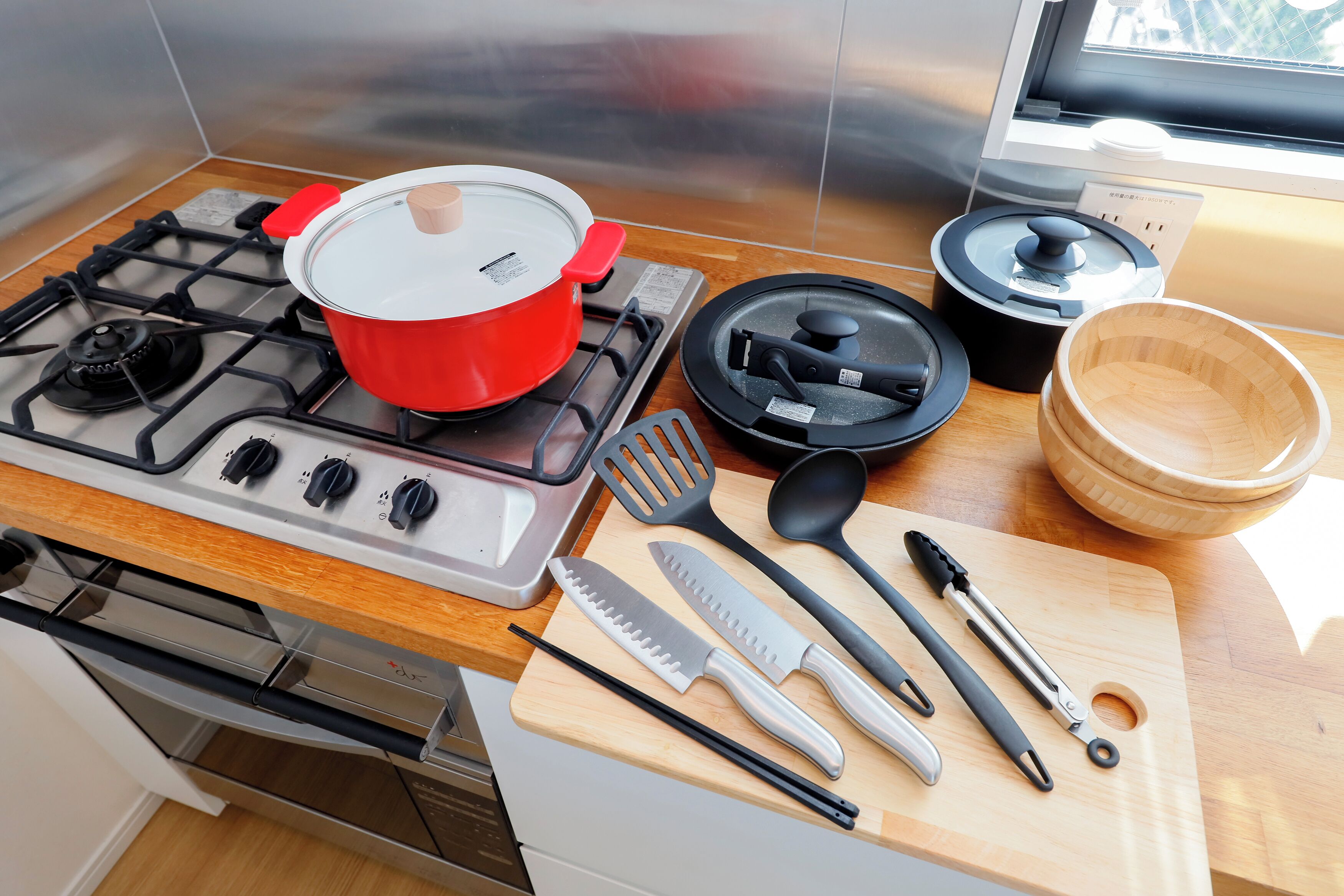 Een keuken met een gasfornuis, rode pan, zwarte koekenpan, houten snijplank en diverse keukengerei.
