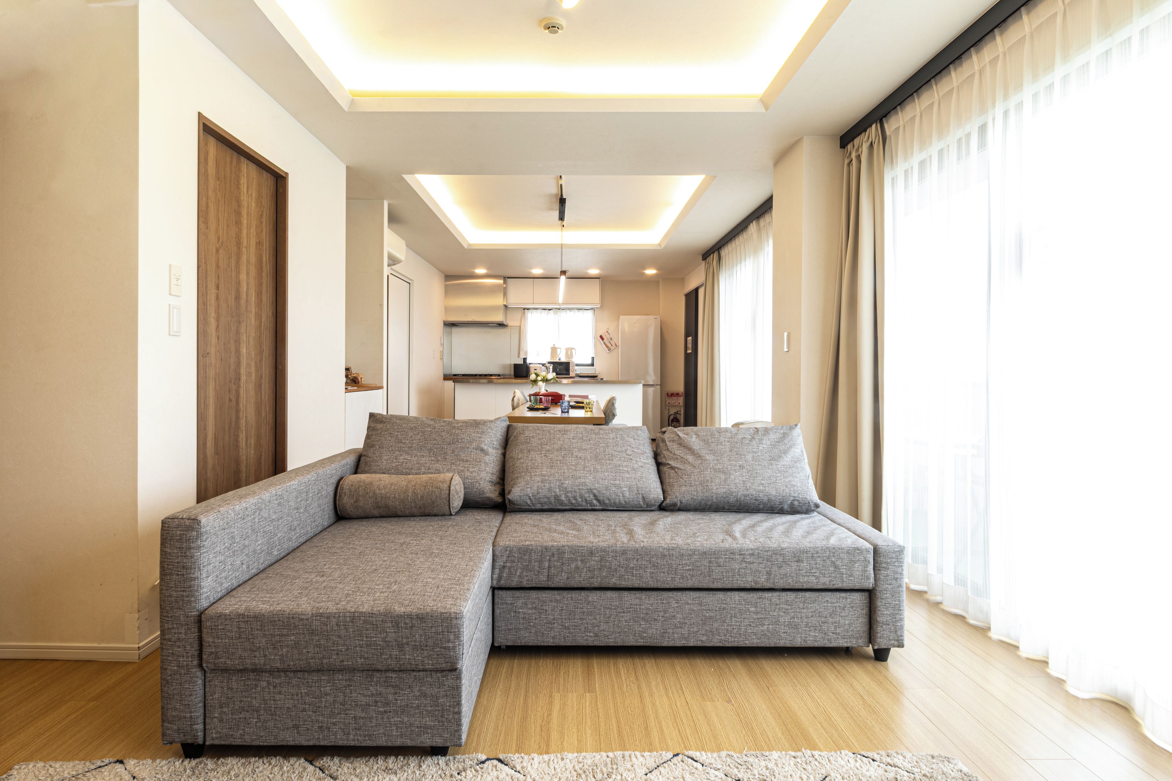 Una sala de estar moderna con un sofá modular, una cocina al fondo y un amplio ventanal con cortinas.