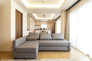 A modern living room with a sectional sofa, a kitchen in the background, and a large window with curtains.