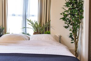 A neatly made bed with a striped bedspread, a potted plant on the windowsill, and a tall plant beside the bed.