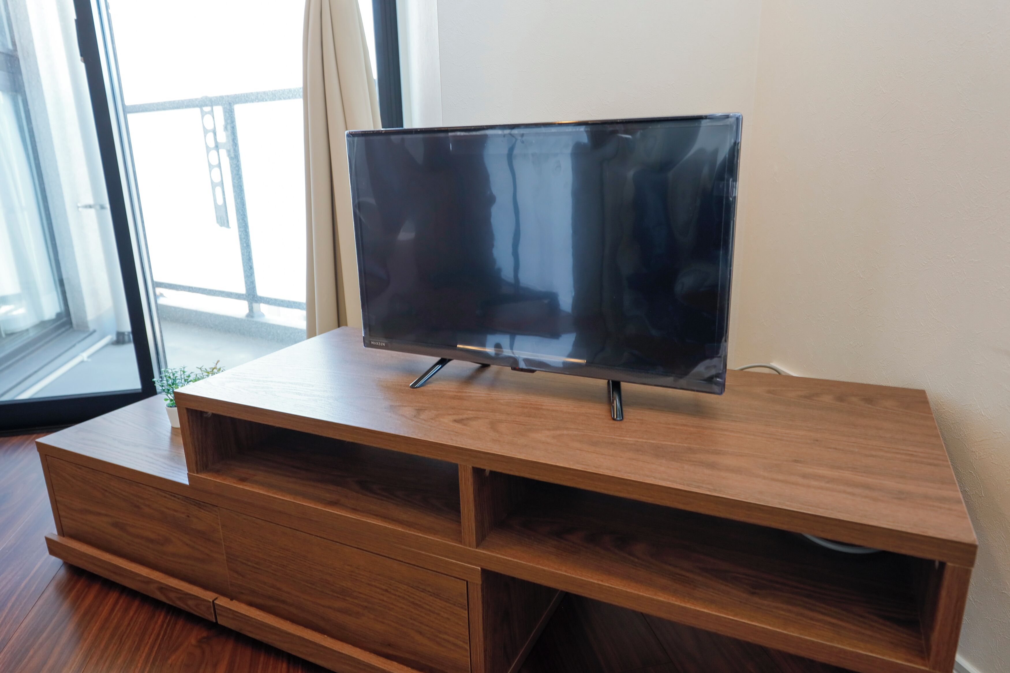 Un meuble en bois pour la télévision, sur lequel est posée une télévision à écran plat, se trouve dans une pièce dotée d’une grande fenêtre et d’un balcon.
