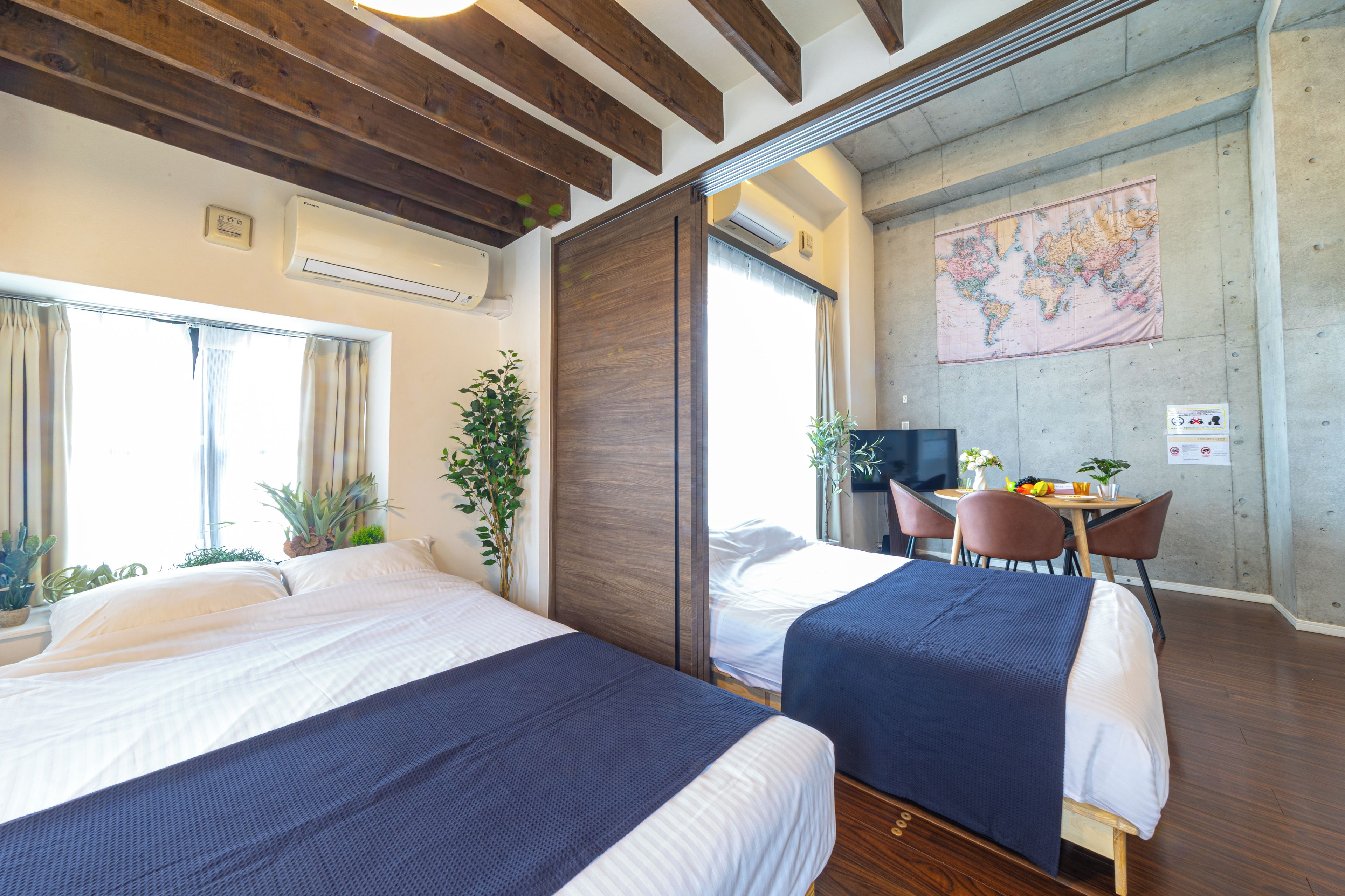 A hotel room with two beds, a desk with a chair, a map on the wall, and a window with curtains.