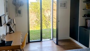 Moderni makuuhuone, jossa on puulattia, sinisellä peitolla varustettu sänky, työpöytä tuolilla ja suuri liukuovi, joka johtaa puutarhaan.