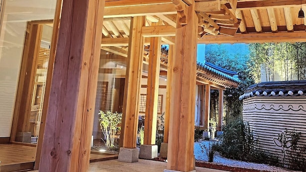 Una estructura de madera de diseño tradicional, posiblemente un pabellón o mirador, con vista a un jardín y un edificio con tejado de baldosas al fondo.