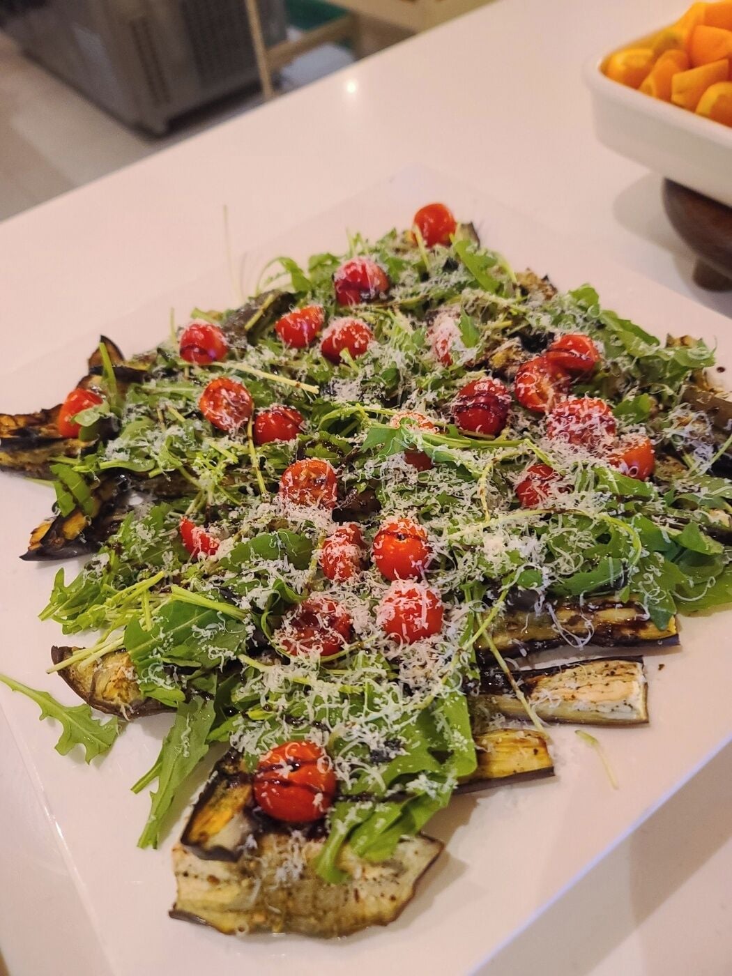 Prato branco retangular com salada de rúcula, tomate cereja e fatias de berinjela grelhada, coberta com queijo ralado.