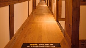 Un pasillo con suelo y paredes de madera que conduce a una puerta con un letrero que dice 'Quítate los zapatos y experimenta hanok'.