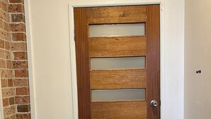 A wooden door with horizontal slats and a metal handle, set in a corner with a brick wall and a potted plant. - Emerald Beach Family Retreat (Emerald Beach)