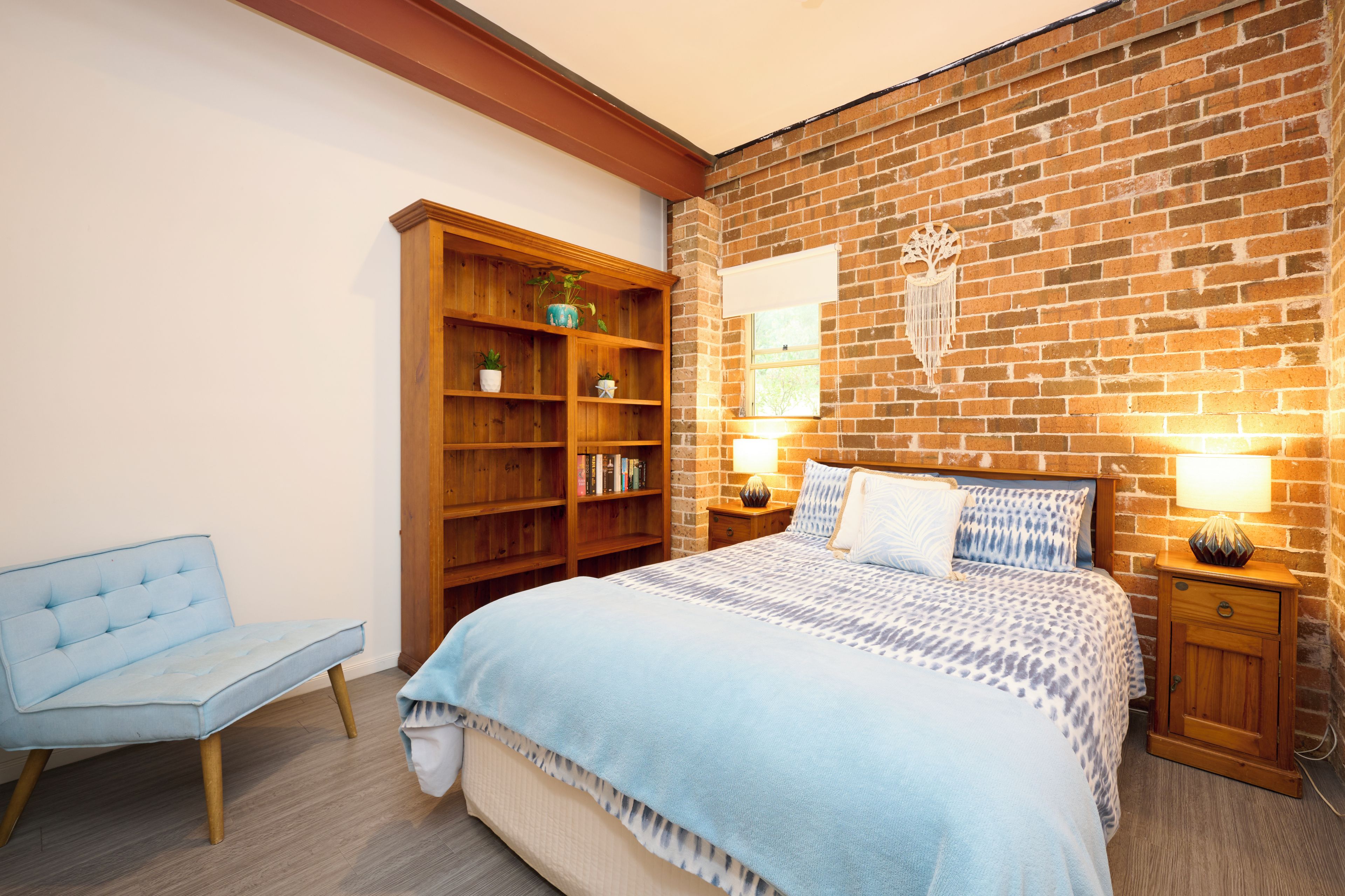 Une chambre avec un mur en briques, un lit avec une literie à motifs, une table de chevet en bois, une lampe sur la table de chevet, une chaise bleue et une bibliothèque en bois avec des plantes.