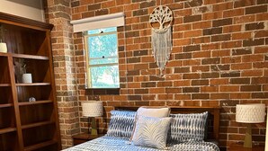A bedroom with a brick wall, a large bed with patterned bedding, two bedside lamps, and a wooden nightstand.
