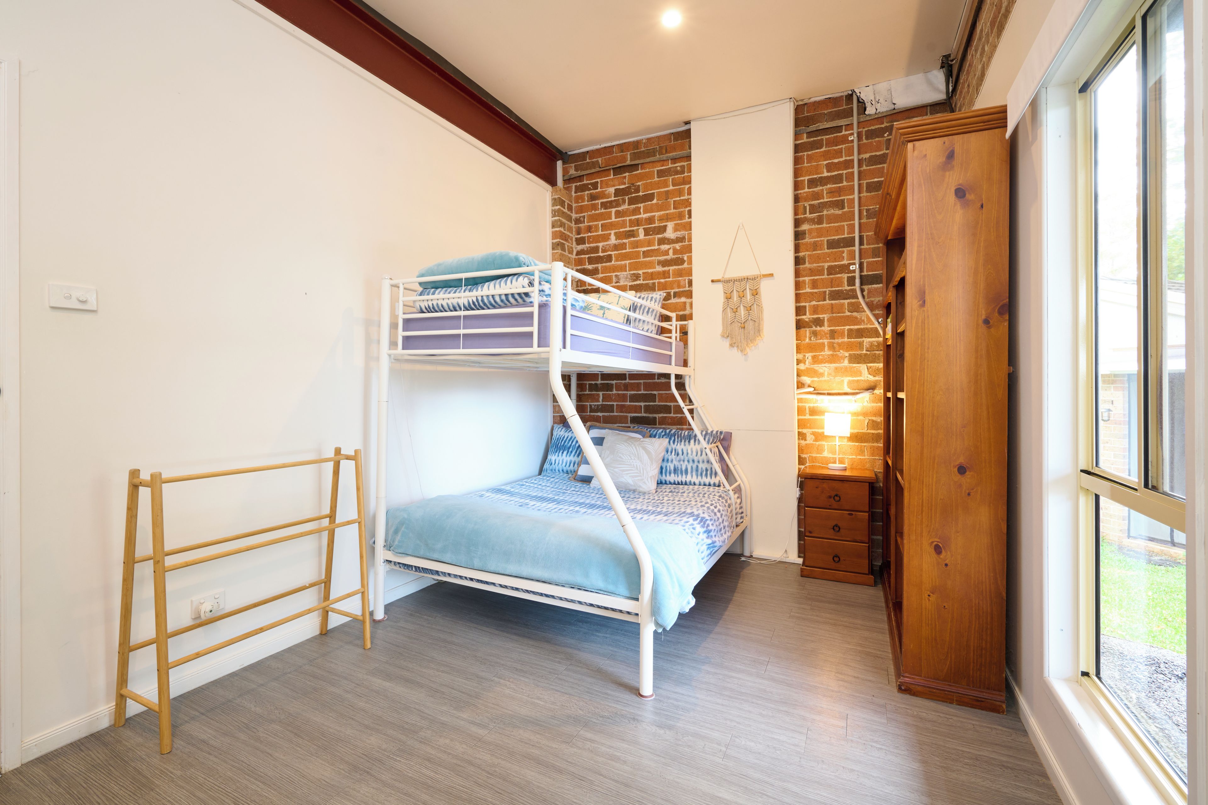 A bunk bed with a wooden ladder, a small wooden dresser, and a wall-mounted macrame decoration in a room with a brick wall and a window.