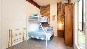 A bunk bed with a wooden ladder, a small wooden dresser, and a wall-mounted macrame decoration in a room with a brick wall and a window.