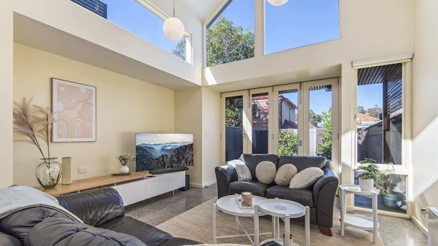 A modern living room with a black leather sofa, a flat-screen TV, and a large window offering a view of the backyard. - Gorgeous 2-Bed Victorian Home in Artsy Suburb (Brunswick)