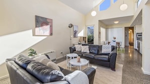 A spacious living area with a black leather sofa, a small round table, and a dining table with chairs. There is a large window providing natural light and a view of the outdoors. - Gorgeous 2-Bed Victorian Home in Artsy Suburb (Brunswick)