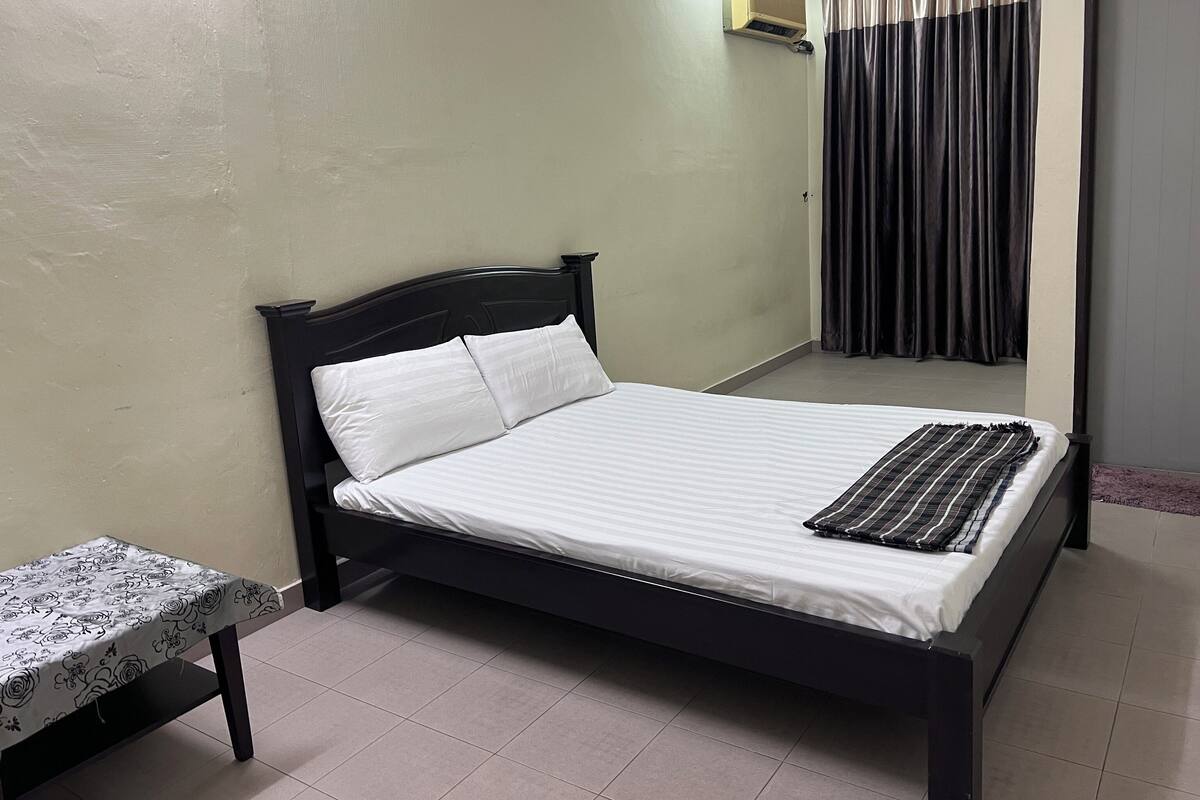 A neatly made bed with white linens and a striped blanket, a small table with a floral tablecloth, and a window with dark curtains.