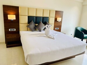 A hotel room with a large bed, a wooden headboard, two wall-mounted lamps, and a green chair. - Mastiff Grand Dharwad (Dharwad)