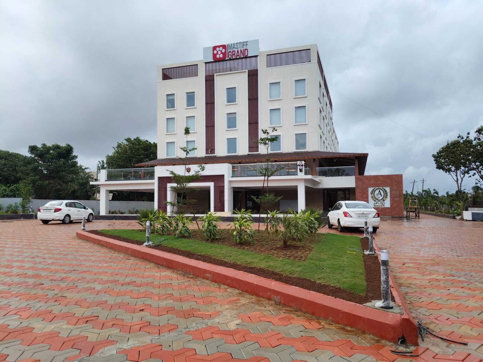 A modern hotel with a prominent IMASTIFF GRAND sign, a covered parking area, and a well-maintained garden.
