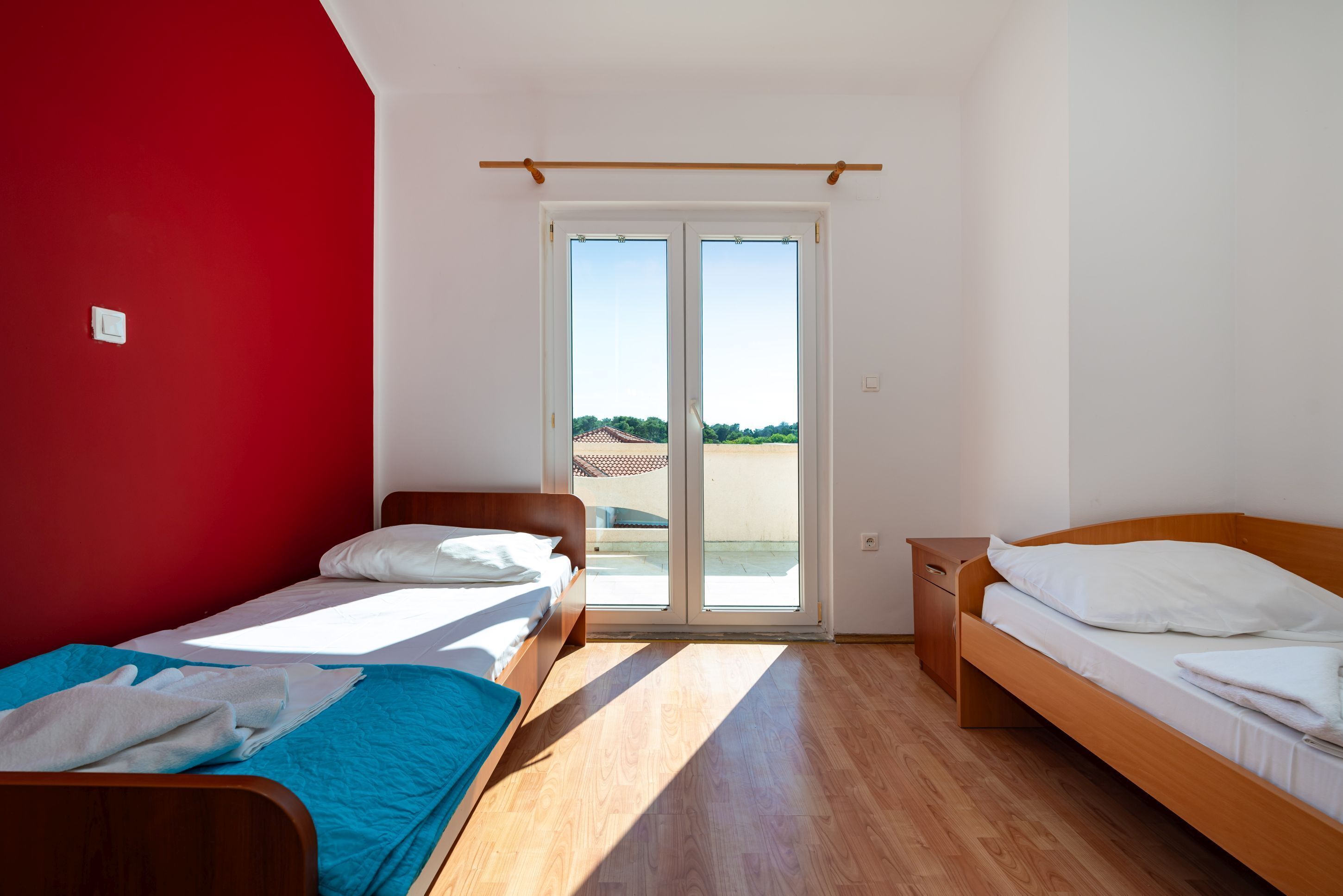 A room with two beds, a red wall, a wooden floor, and a sliding glass door leading to an outdoor area.