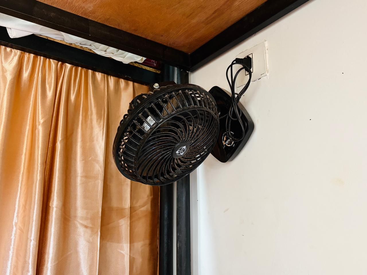 Un ventilador de pared negro en una esquina, con una cortina cubriendo parcialmente una ventana.