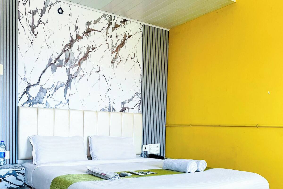 A modern hotel room with a marble-patterned headboard, a large bed with white linens, a green throw blanket, and a yellow accent wall.