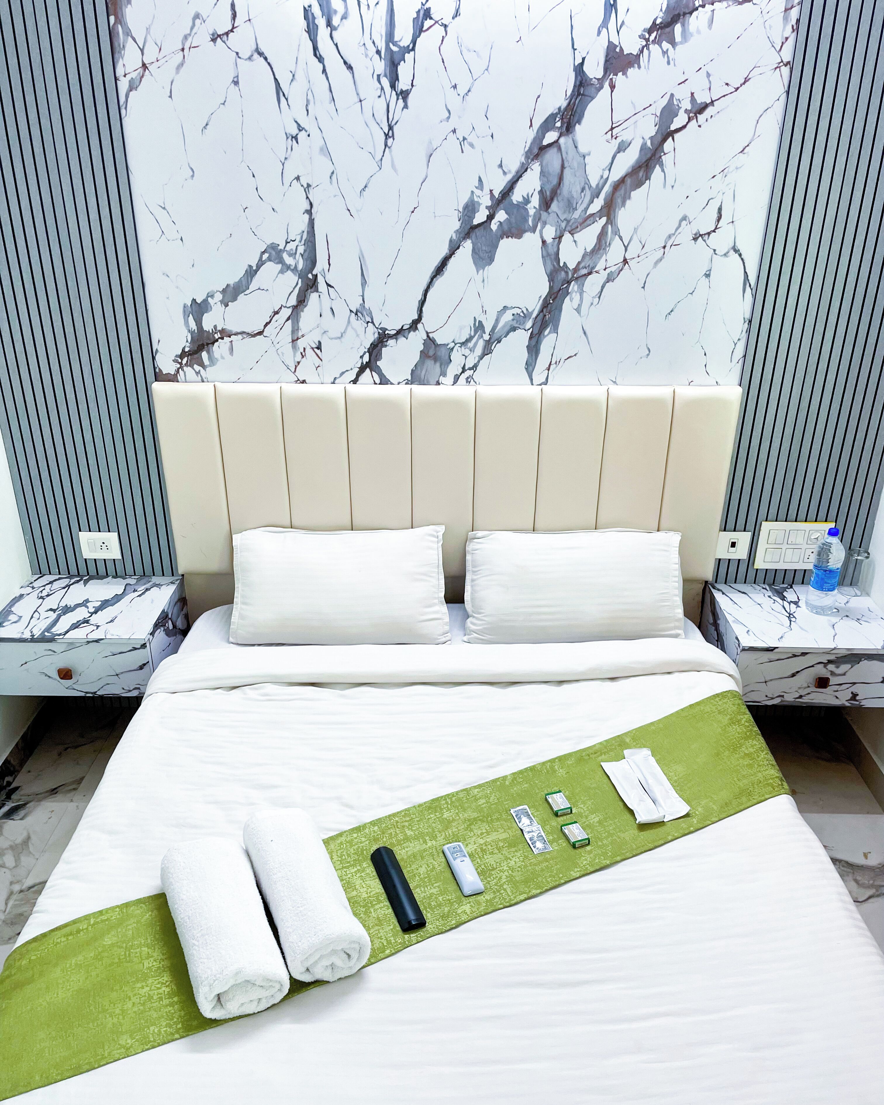 A hotel room with a marble-patterned headboard, a neatly made bed with white linens, a green towel, and a remote control on a green mat.