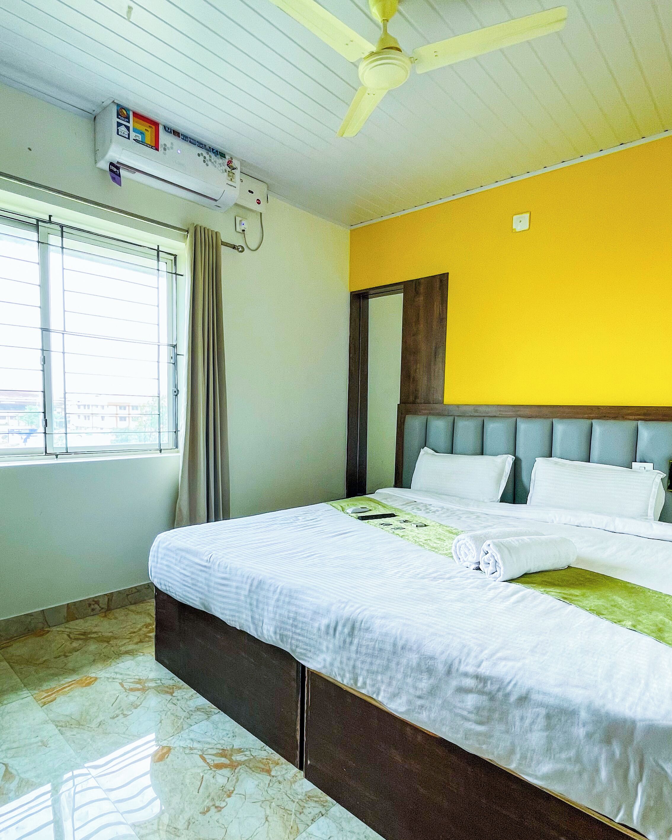 A hotel room with a bed, a ceiling fan, an air conditioning unit, a window with curtains, and a yellow wall.